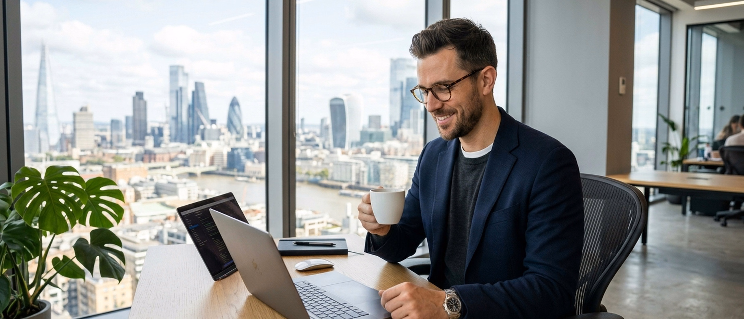 A relaxed professional in glasses looking at his laptop with a cup of coffee at a high-rise office desk overlooking the city skyline.